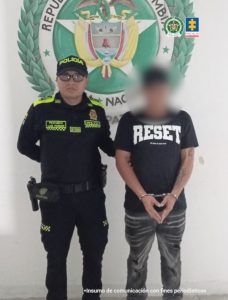 En la imagen se observa a un agente de la Policía Nacional de Colombia (identificable por su uniforme oficial y gorra con la palabra "POLICÍA") posando junto a un hombre esposado, cuya identidad ha sido difuminada o desenfocada para proteger su privacidad.Ambos están de pie frente a una pared blanca con el escudo de la República de Colombia y los logotipos de la Policía Nacional y la Fiscalía General de la Nación, lo que sugiere que se trata de una captura oficial. El detenido lleva una camiseta negra con la palabra "RESET" en letras grandes blancas, y debajo una frase más pequeña que dice “It’s time to start over”. Sus manos están esposadas al frente, lo cual indica que ha sido retenido formalmente por las autoridades. En la parte inferior de la imagen se aclara: "Insumo de comunicación con fines periodísticos".