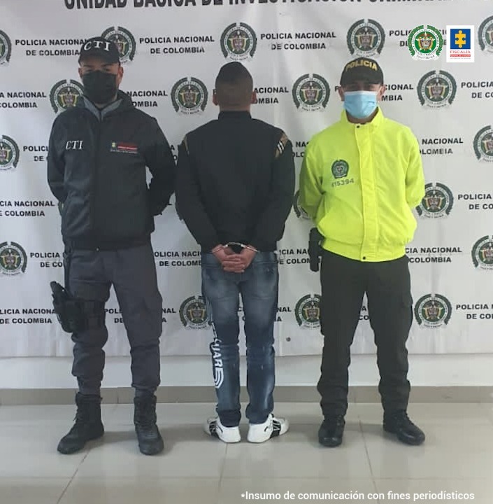 En la fotografía se observan tres personas, una de ellas el capturado, un uniformado de la Policía Nacional y un funcionario del CTI. En la parte posterior se observa un backing con escudos de la Policía Nacional.