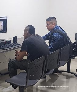 En la fotografía se aprecia a Cristhian Alexis Rivas Sánchez sentado, dentro de una habitación, sentado en unas sillas negras y frente a un computador. Viste camiseta y pantalón negro, así como zapatillas blancas. A su lado izquierdo hay un servidor del Inpec.