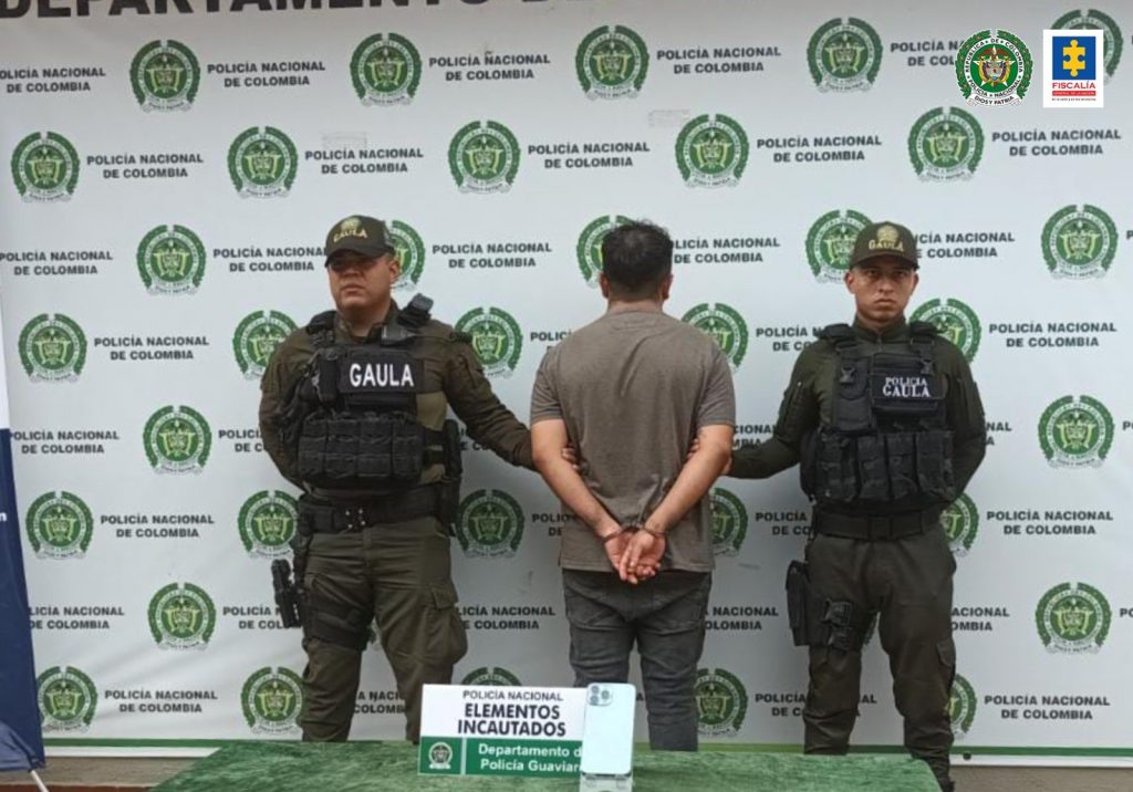 Está el capturado de espaldas esposado, custodiado por dos uniformados de la Policía. Delante una mesa con material incautado. Detrás Banner de la Policía Nacional.