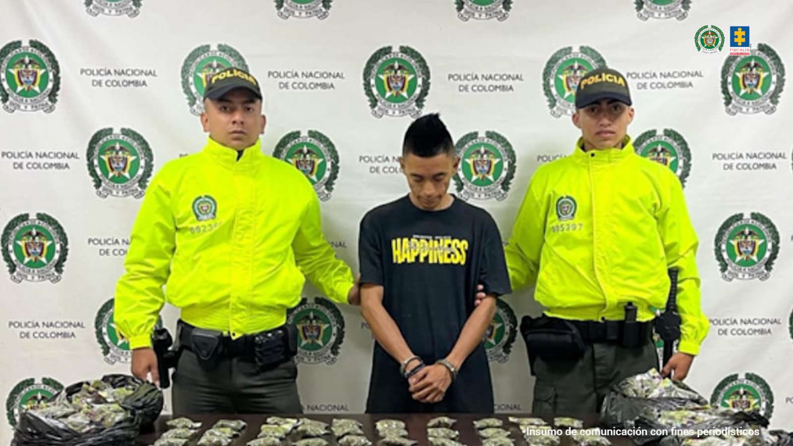 En la fotografía se aprecia de pie, con los brazos adelante y esposado a Cristian Andrés Alzate, alias Cara de gallo. Viste camiseta de color negro, demanga corta y con estampado amarillo a la altura del pecho. A sus extremos hay uniformados de la Policía Nacional. Delante de ellos hay una superficie de color marrón y sobre ella gran cantidad de bolsas plásticas con contendio vegetal. Asimismo, un letrero que dice ‘Marihuna’. Detrás de ellos hay un pendón institucional de la Policía.