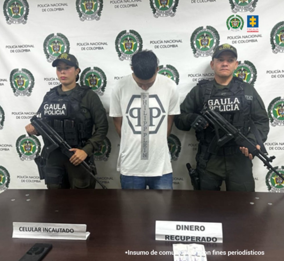 En la fotografía se aprecia de pie, con la cabeza agachada y con los brazos atrás a Cristian Andrés Carmona Marín. Viste camiseta blanca, de manga corta, con estampado en forma de rombo y en el centro de éste dos letras ‘P’; asimismo, usa jean azul. A sus extremos hay uniformados de la Policía Nacional. Delante de ellos hay una superficie de color marrón y sobre ella hay dos letreros que dicen: ‘Celular incautado’ y ‘Dinero recuperado’. Detrás de ellos hay un pendón institucional de la Policía Nacional.