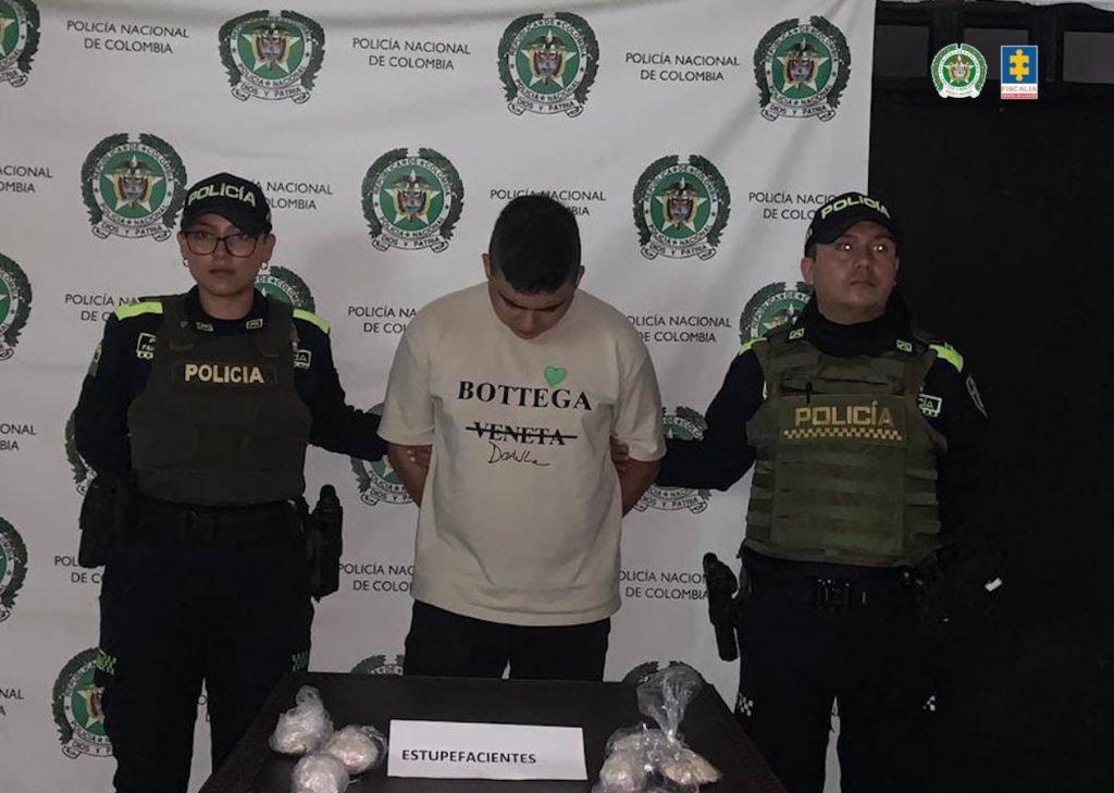 En la fotografía se aprecia de pie, con los brazos atrás y la cabeza agachada a Cristinan Andrés Giraldo Zapata. Viste camiseta de manga corta, de color beage y a la altura del pecho lleva impresas, en negro, las palabras ‘Bottega’ y ‘Veneta’; asimismo, pantalón negro. A sus extremos hay uniformados de la Policía Nacional. Delante de ellos hay una superficie y sobre ella hay seis paquetes en bolsas plásticas con contenido blanco. Asimismo, un letrero que dice: ‘Estupefacientes’. Detrás de ellos hay un pendón institucional de la Policía.