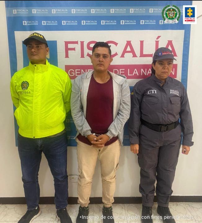 capturado de frentes esposado manos adelante, custodiado por investigadora CTI de la Fiscalía y uniformado de la Policía. Detrás banner de la Fiscalía.