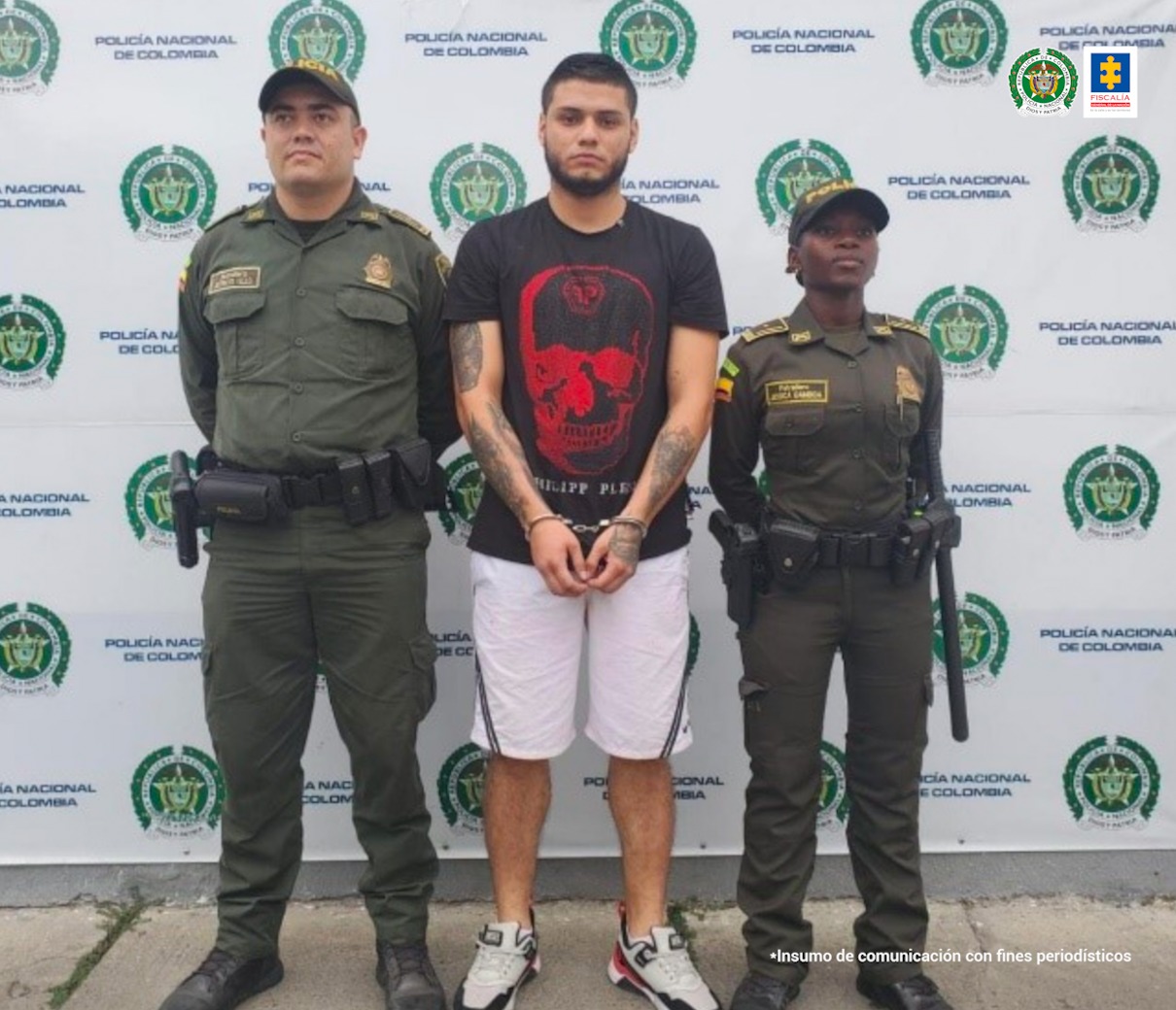 En la foto aparece el judicializado vestido con camiseta negra negra con estampado rojo, pantaloneta blanca y tenis. A lado y lado de él aparecen dos integrantes de la Policía y detrás de ellos el pendón institucional de la Policía Nacional.
