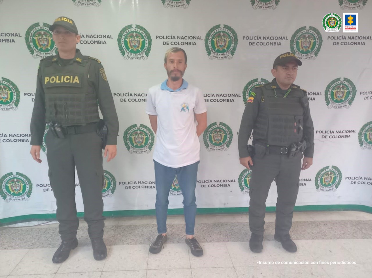 En la foto aparece, de pie y con los brazos atrás, Cristian Augusto Colorado Caro. Vista camiseta blanca de manga corta, jean azul y zapatos oscuros. A lado y lado de él hay uniformados de la Policía Nacional. Detrás de ellos hay un pendón institucional de la Policía.