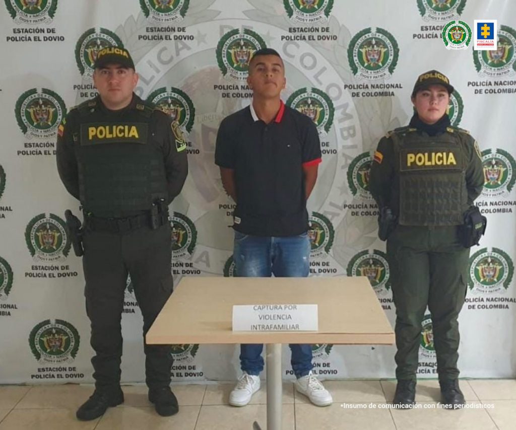 En la foto aparece, de pie y con los brazos atrás Cristian Camilo Camacho Guazaquillo. Viste camisa manga corta y con cuello color negra con apliques blancos y rojos; jean azul y zapatillas blancas. A lado y lado de él hay dos uniformados de la Policía Nacional. Delante de ellos hay una mesa cuadrada y detrás de ellos hay un pendón institucional de la Policía.