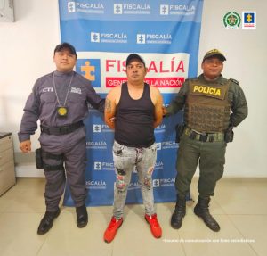 En la fotografía aparece un capturado, junto a personal de Fiscalía General de la Nación y Policía Nacional. En la parte posterior de la imagen se ven banners de Policía Nacional y la Fiscalía General de la Nación.