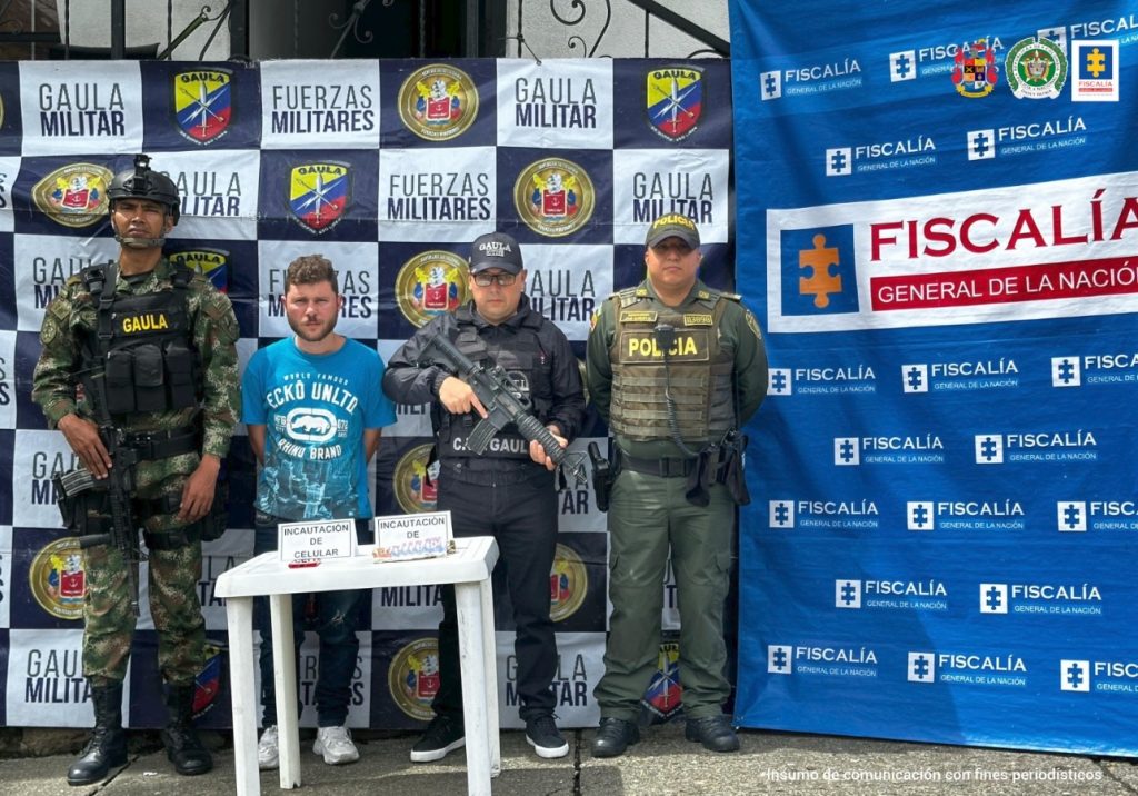 Se observa al capturado y a cada costado funcionarios de Policía Nacional, Ejército nacional. Atrás un pendón de Fiscalía general de la Nación y de Policía Nacional