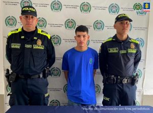 Cristian Camilo Mena Díaz tiene a los lados dos policías y en el fondo se ve un pendón con logos de la institución.