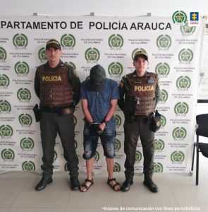 En la imagen aparece dos uniformados de la Policía Nacional con un capturado. En la parte posterior se observa el banner de la Policía Nacional del departamento de Arauca
