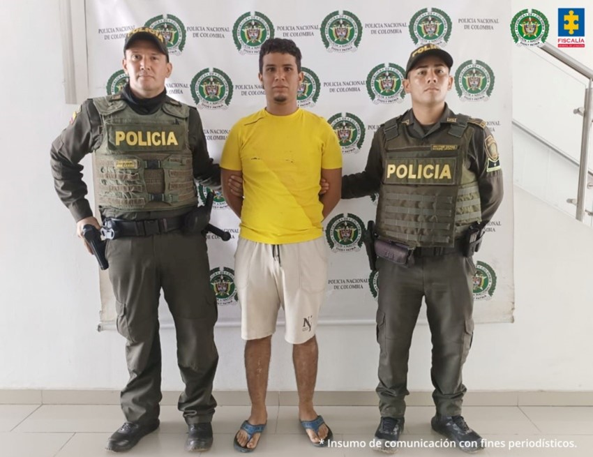 Se visualiza al capturado junto a dos uniformados de la Policía Detrás se ubica el banner que identifica a la Policía Nacional.