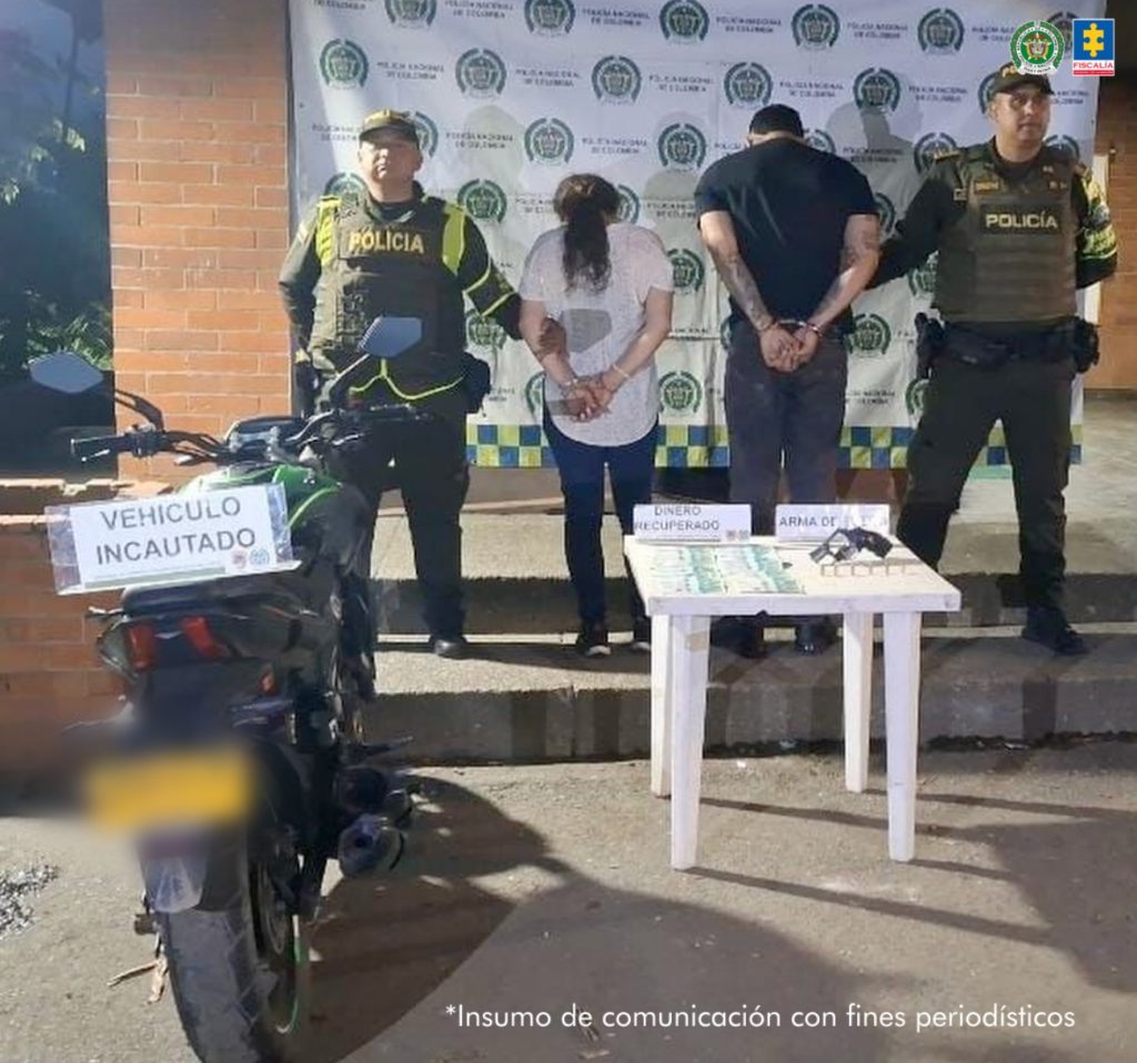En la fotografía se aprecian de pie, con los brazos atrás, esposados y de espaldas a la cámara a Cristian Joan Ríos Castellanos y Judith Lorena Rojas Muñoz. La mujer viste con blusa de color rodado, jean azul oscuro y zapatillas negras. El hombre viste camisa de color negro y manga corta, así como jean azul oscuro. A los extremos hay uniformados de la Policía Nacional. En la parte delantera hay una mesa blanca cuadrada y sobre ella se ve dinero, un arma de fuego y munición. Sobre la misma superficie hay dos letreros que dicen: ‘Dinero recuperado’ y ‘Arma de fuego’. Asimismo, al lado izquierdo de la fotografía hay una motocicleta y sobre ella un letrero que dice: ‘Vehículo incautado’. En la parte posterior, hay un pendón institucional de la Policía.
