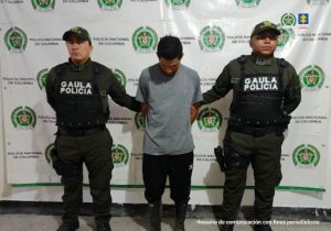 El capturado se encuentra de frente y custodiado por dos uniformados del Gaula de la Policía Nacional. Tiene unas botas de caucho, un buso gris y un pantalon negro.