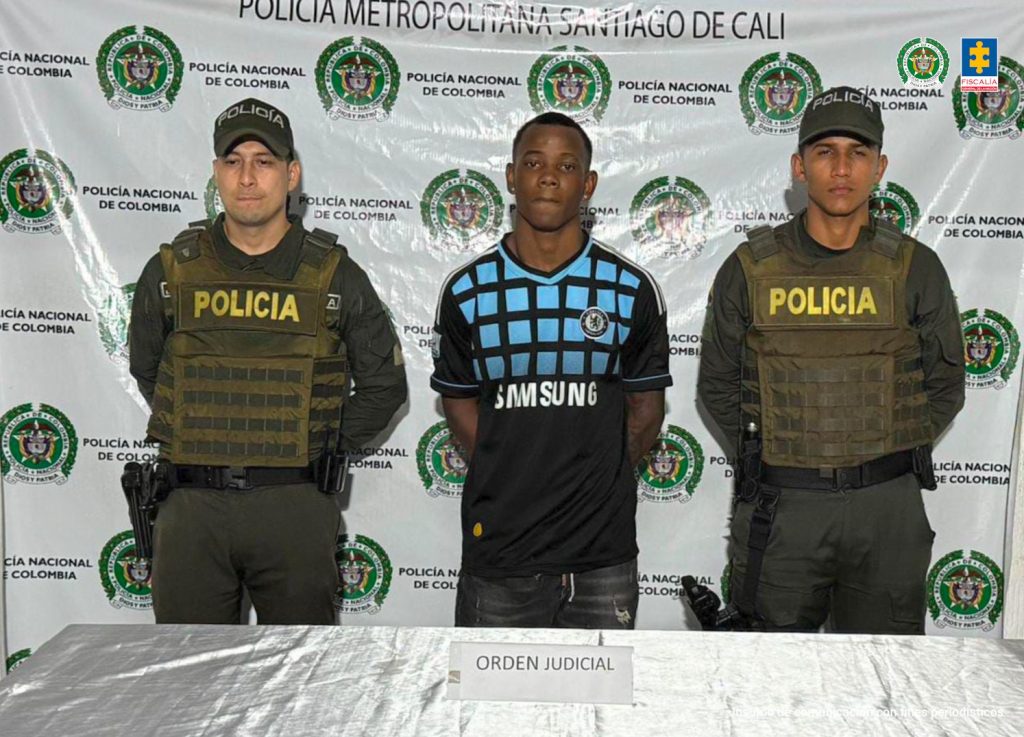 En la fotografía aparece de pie y con los brazos atrás Cristhoper Victoria Trujillo. Usa aretes (topos) en ambas orejas. Viste con camiseta de color negro, mangas cortas y estampados cuadrados de color azul en la parte del pecho; jean de color gris oscuro desgastado. En sus extremos hay uniformados de la Policía Nacional. Delante de ellos hay una superficie y sobre ella hay un letrero blanco que dice: ‘Orden judicial’. Detrás de ellos hay un pendón institucional de la Policía.