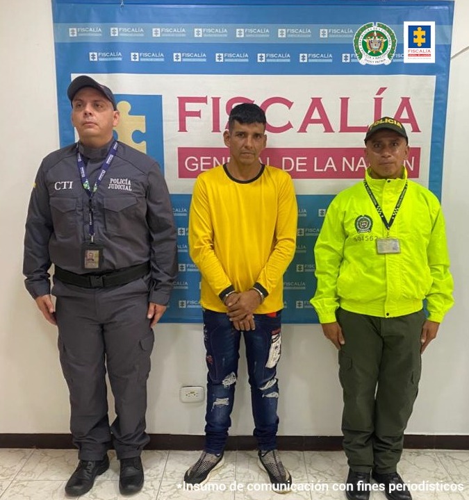 capturado y esposado de frente custodiado por investigador del CTI de la Fiscalía e integrante de la Policía. Banner de la Fiscalía.
