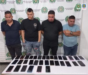 En la foto se aprecia de pie a cuatro hombres. Tres de ellos visten camiseta negra de manga corta, pantalón negro y jean azul. Otro viste camiseta azul clara de manga corta y jean azul. Delante de ellos hay una superficie blanca y sobre ella gran cantidad de teléfonos celulares. Detrás de ellos hay un pendón institucional de la Policía Nacional.