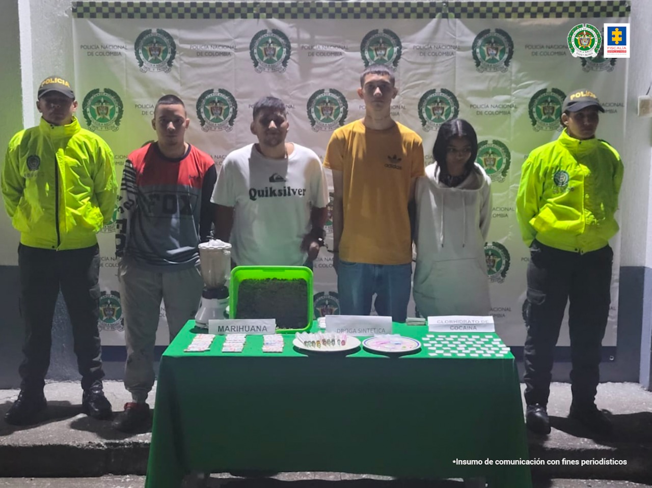 En la foto aparecen de pie los cuatro presuntos implicados en la conservación de estupefacientes. Hay tres hombres y una mujer. A lado y lado de ellos hay uniformados de la Policía Nacional. Delante de ellos hay una mesa con mantel verde y sobre ella hay una licuadora, jeringas y papeletas. También hay dos letreros que dicen ‘marihuana’ y ‘droga sintética’. Detrás de ellos hay un pendón institucional de la Policía.