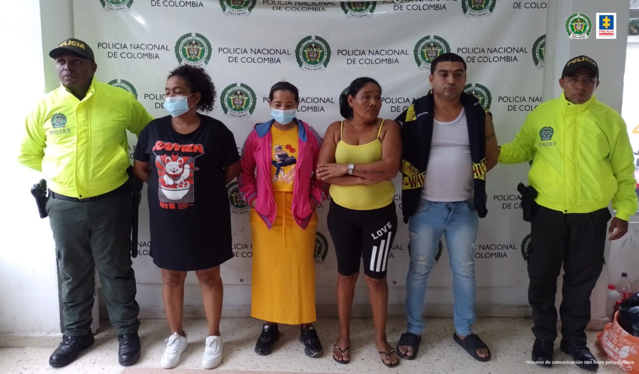 Los cuatro detenidos se encuentran acompañados de dos uniformados de la Policía Nacional. De fondo está un pendón de la Policía Nacional con el logo oficial.
