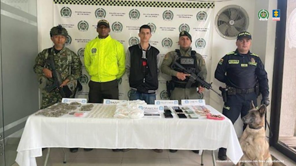 Se observa al capturado de frente y a cada costado dos funcionarios de Policía Nacional que lo custodian, atrás, un pendón de Policía Nacional. A un costado un canino de Policía Nacional. Adelante una mesa en la cual están expuestos los elementos incautados en la diligencia de allanamiento