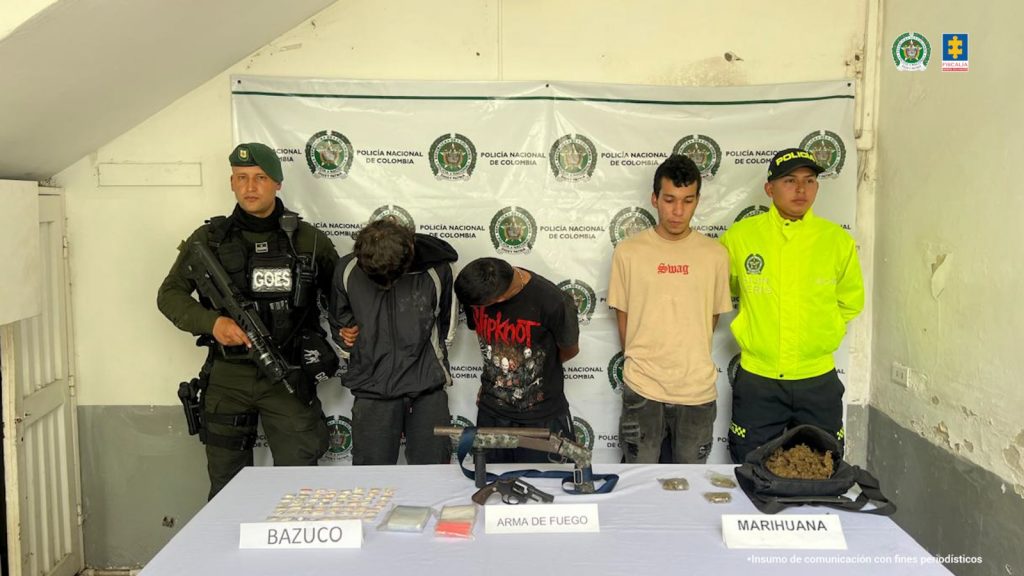 Se observan tres capturados de frente, en fila. A cada costado un funcionario de Policía Nacional que los custodian. Atrás un pendón de Policía Nacional. Adelante una mesa en la cual están expuestos los elementos incautados en la diligencia de allanamiento