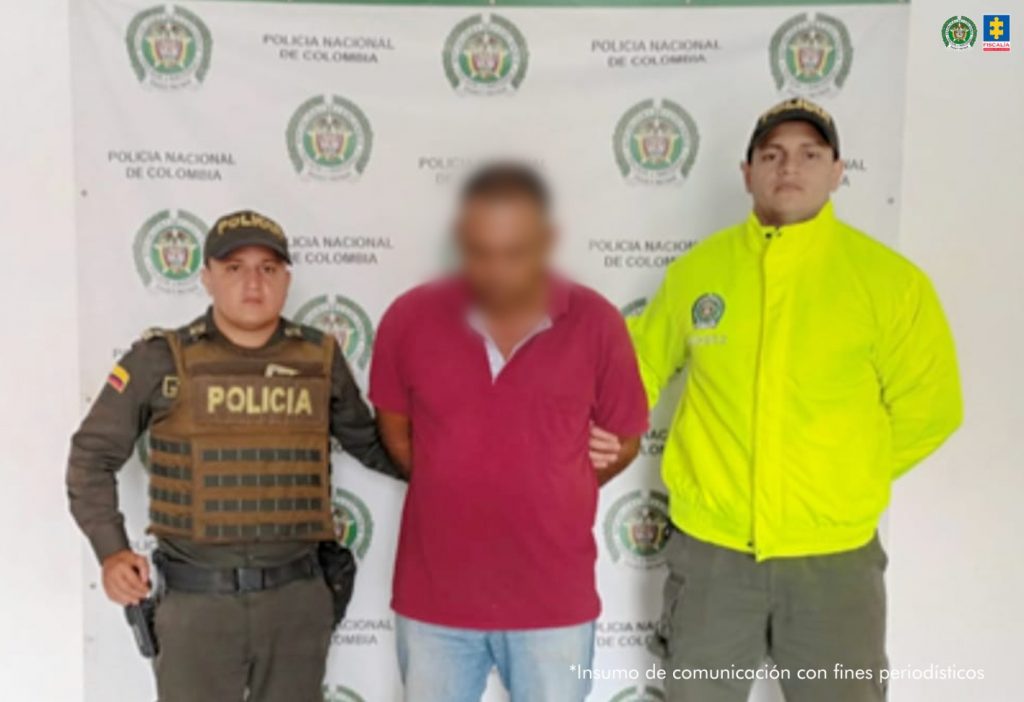 En la fotografía se aprecia de pie y con los brazos atrás al investigado por acceso carnal violento agravado. Viste camiseta de color rojo,manga corta y jean azul claro. A los extremos de él hay uniformados de la Policía Nacional. Detrás de ellos hay un pendón institucional de la Policía.