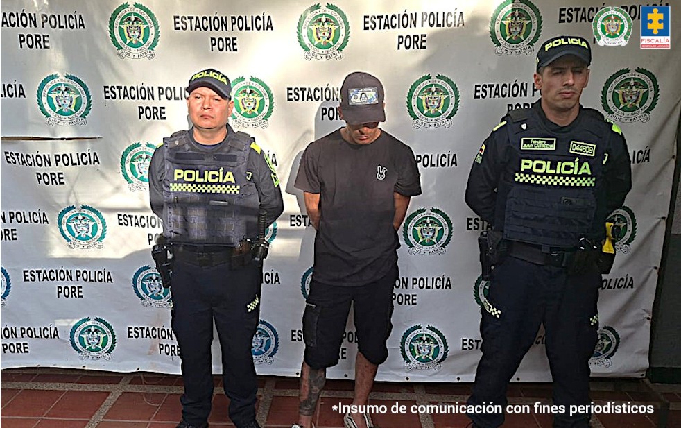El capturado aparece con sus manos esposadas a la espalda, tiene una gorra y viste una camiseta y short negros, está custodiado por uniformados de la Policía.