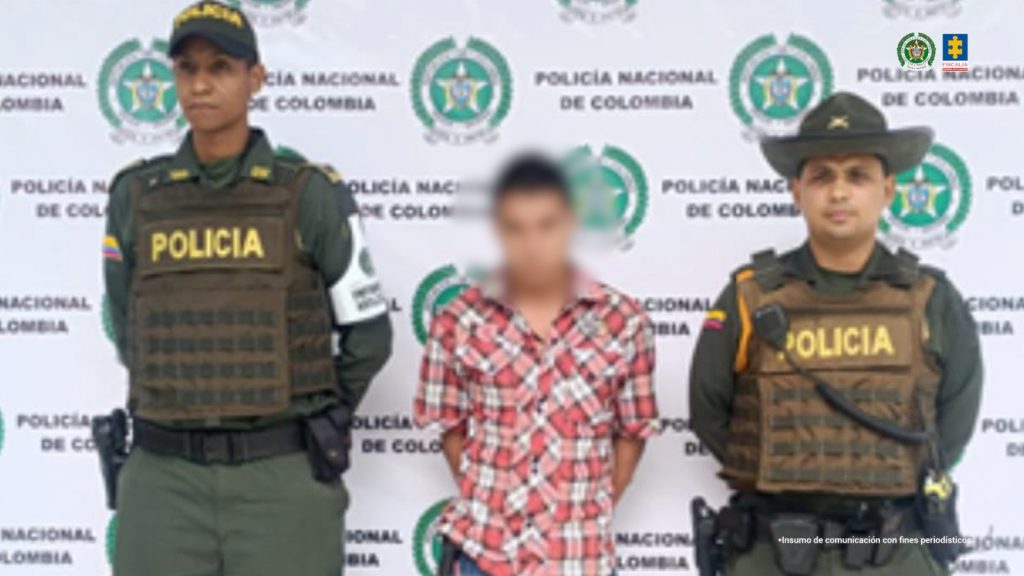 En la fotografía se aprecia de pie y con los brazos atrás al imputado. Viste camisa de cuadros rojos y blancos, jean azul desgastado y zapatos azules. A sus extremos hay uniformados de la Policía Nacional y detrás de ellos hay un pendón institucional de la Policía.