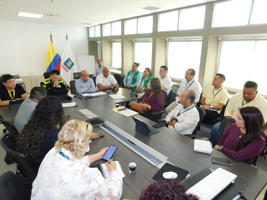 Reunión de mesa técnica interinstitucional en una sala de juntas, con la participación de autoridades y funcionarios sentados alrededor de una mesa. En las imágenes se observan representantes de la Policía, Fiscalía, Defensoría del Pueblo, ICBF y otras entidades escuchando y tomando notas durante la discusión. Al fondo se ven las banderas de Colombia y de la Fiscalía, mientras varios asistentes intervienen y otros siguen atentos la conversación.