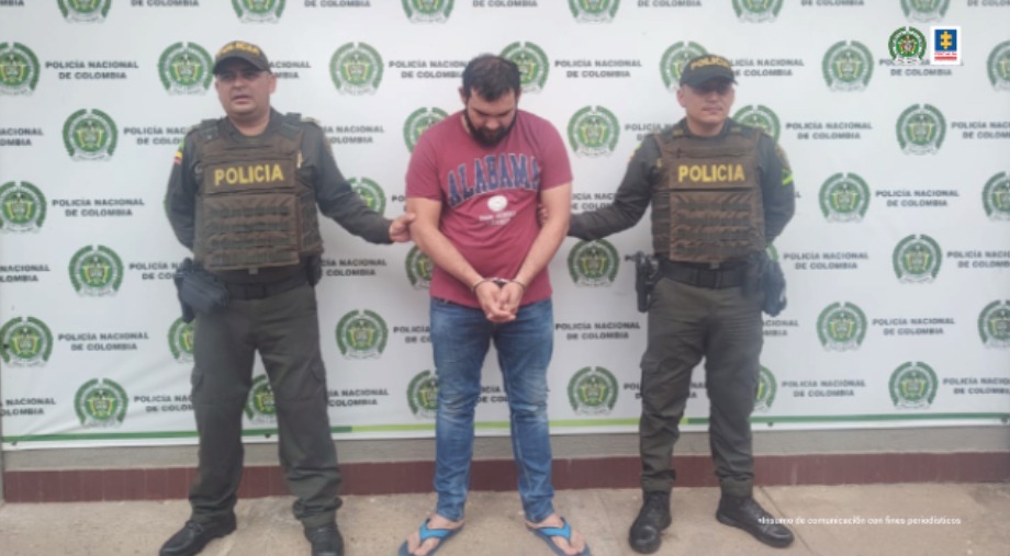 Está el capturado de frente, cabeza agachada y manos esposadas adelante, custodiado por dos uniformados de la Policía. Detrás banner de la Policía.