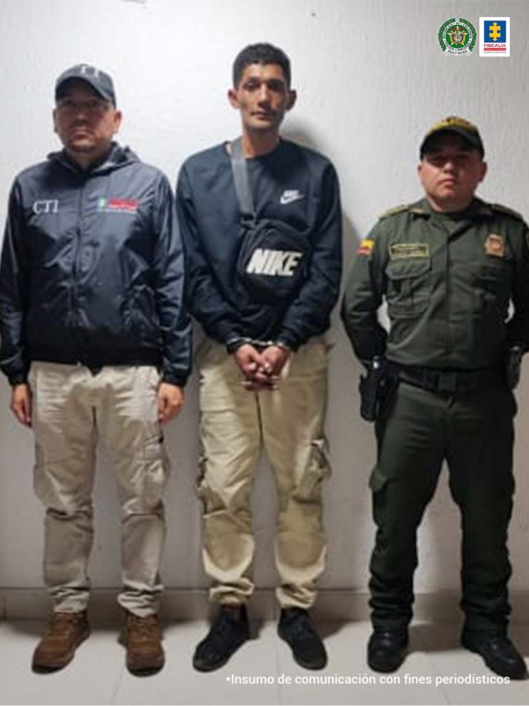 En la foto aparece Daniel Armando Velasco Villamil, con los brazos adelante y esposado; viste buzo de manga larga oscuro, pantalón largo caqui, zapatos negros y carga un canguro oscuro. Al lado derecho de él hay un servidor del CTI de la Fiscalía y a la izquierda un uniformado de la Policía Nacional.