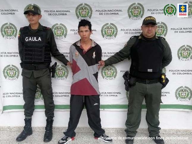 El hoy imputado aparece vestido con camiseta estampada con colores rojo, gris negro y un pantalón tipo sudadera negro y tenis. A lado y lado de él aparecen dos integrantes del Gaula de la Policía Nacional y detrás de ellos aparece un pendón institucional de la Policía.