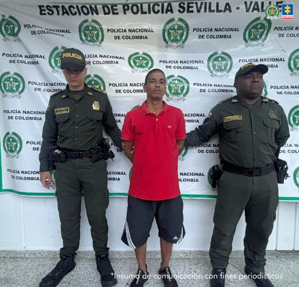 En la fotografía de pie y con los brazos atrás Daniel Núñez Tamayo. Viste con camisa de manga corta, de color rojo, bermuda de color gris oscuro con rayas blancas verticales en la parte inferior y a los lados; usa zapatillas negras. A sus extremos uniformados de la Policía Nacional. Detrás de ellos hay un pendón institucional de la Policía.