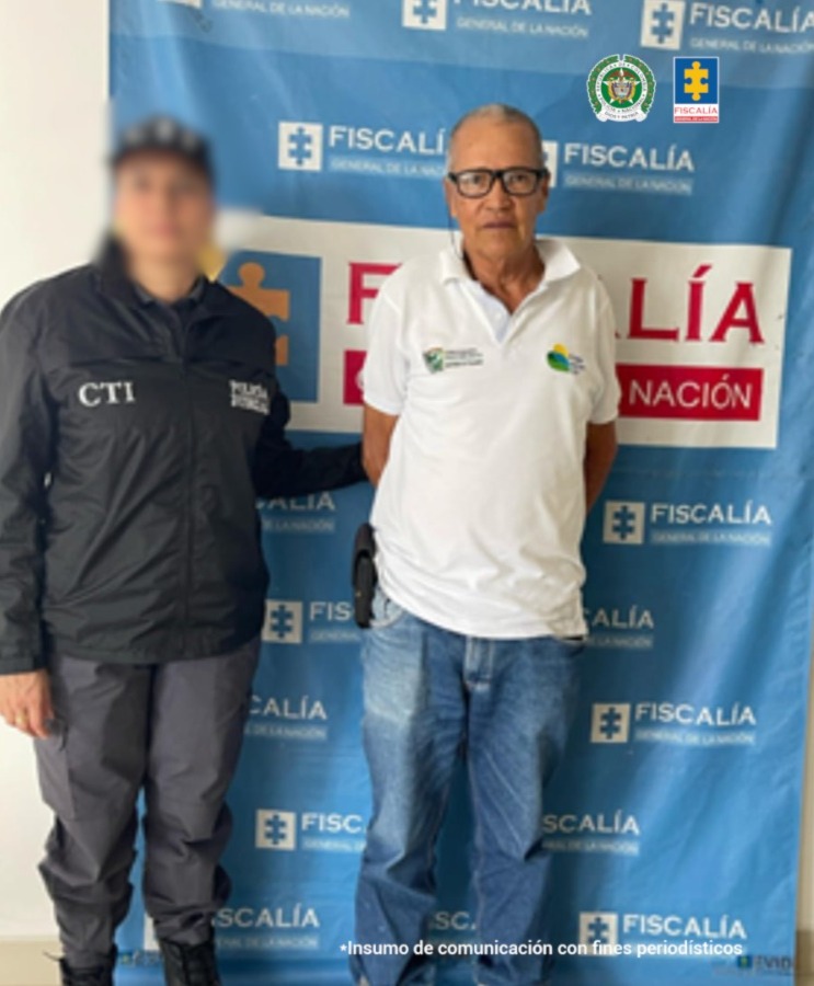En la fotografía se aprecia de pie y con los brazos atrás a Darío Arnulfo Morales Franco. Viste camisa blanca, jean azul y tenis grises. usa lentes de marco negro. A su derecha hay una servidora del CTI de la Fiscalía y detrás de ellos hay un pendón institucional de la Fiscalía.