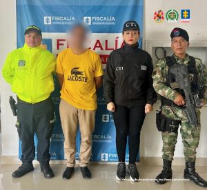 En la fotografía aparece de pie y los brazos atrás Darwin De Jesús Sánchez Palacio. Viste camiseta de manga corta amarilla con la imagen de con caimán y la palabra ‘Lacoste’ en negro; pantalón color caqui y zapatos negros. Al lado izquierdo de él hay una servidora del CTI y un uniformado del Ejército. Al lado derecho hay un uniformado de la Policía Nacional. Detrás de ellos hay un pendón institucional de la Fiscalía.