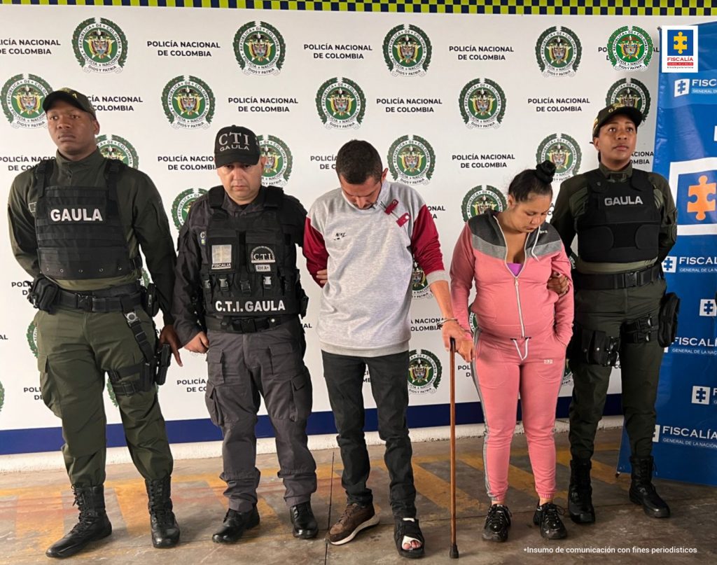 Se observan dos detenidos, un hombre y una mujer. a un costado dos funcionarios de Policía Nacional y al otro uno. Al fondo un pendón de Policía Nacional y uno de Fiscalía General de la Nación.