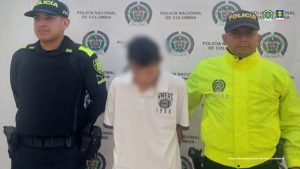 En la fotografía se aprecia de pie y con los brazos atrás a Edier Arturo Muñoz Osorio. Viste camiseta esqueleto de color blanco, bermudas de jean de color azul. Usa lentes de marco negro. A sus extremos hay uniformados de la Policía Nacional.