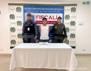 En la fotografía aparece de pie y con los brazos atrás Davinson Ramírez León. Viste con camiseta esqueleto de color blanco y pantalón oscuro. En su extremo derecho hay un servidor del CTI de la Fiscalía y en el izquierdo un uniformado de la Policía Nacional. Delante de ellos hay una mesa con mantel blanco y sobre ella hay un letrero que dice: ‘Capturado Homicidio’. Detrás de ellos hay un pendón institucional de la Policía.