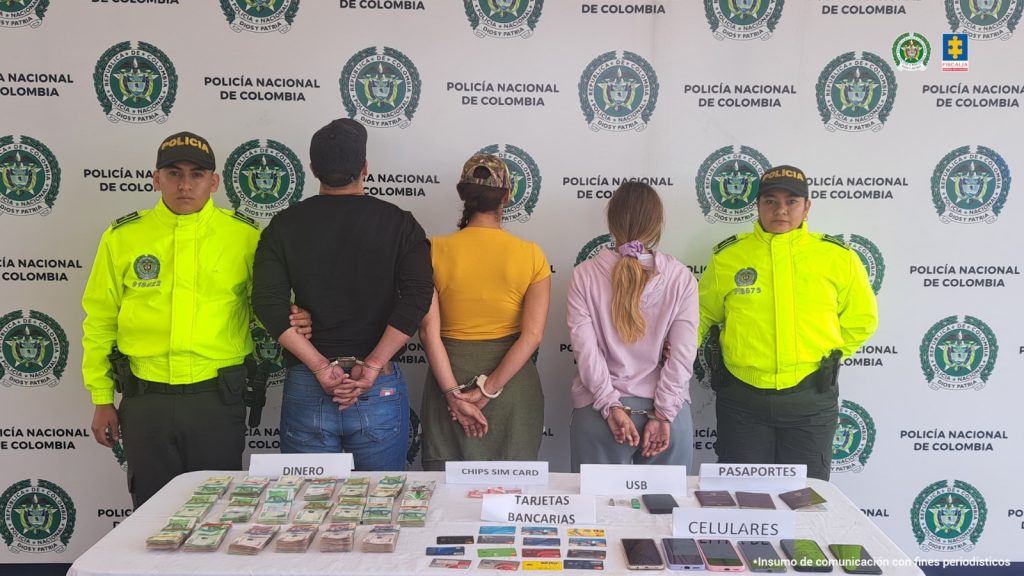 En la imagen están los 3 capturados con Policías a los lados, en la parte de atrás un pendón con el logo de la Policía Nacional en mosaico y adelante una mesa con el dinero y demás elementos incautados