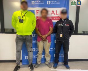  Se visualiza al capturado junto con un investigador del CTI y uniformado de la Policía Nacional, todos con el rostro difuminado. Detrás de ellos el banner que identifica a la Fiscalía General de la Nación.