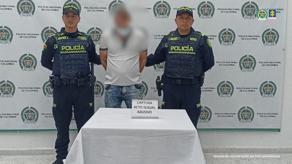Se observa al capturado de frente y sobre su rostro un efecto que lo difumina. A cada costado un funcionario de la Policía Nacional. Atrás, un pendón de Policía Nacional. Adelante, una mesa con el cartel: CAPTURA ACTO SEXUAL ABUSIVO