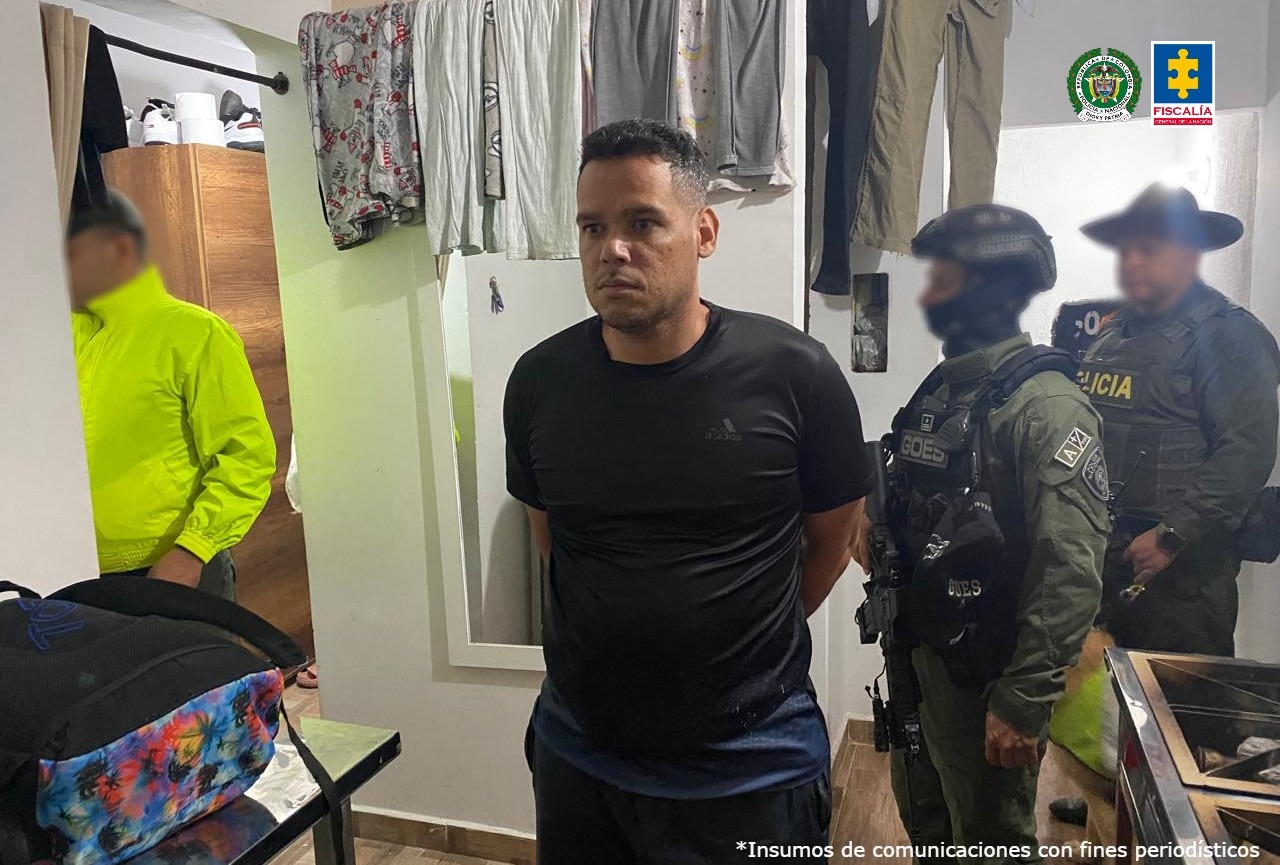 El hombre se ve en el centro de la imagen, dentro de una vivienda. Al fondo puede verse ropa colgada y une spejo, además de miembros del Goes y la Policía. Está esposado, con las manos atrás.