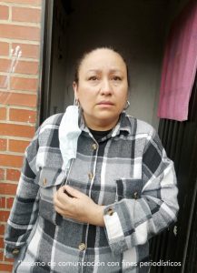 La mujer aparece en el centro de la imagen, de medio cuerpo, con un brazo sobre su estómago. Al fondo se ve una pared de ladrillo visible.