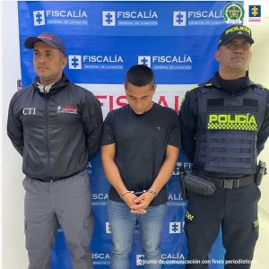 capturado de frente custodiada por investigador del CTI de la Fiscalía y por un policía. Detrás banner de la Fiscalía.