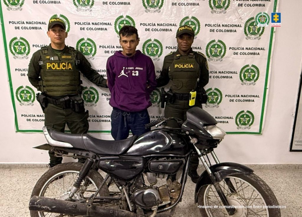 En la fotografía aparece de pie y con los brazos atrás. Viste con buso de color vinotinto con estampado blando en el pecho y pantalón negro. En sus extremos hay uniformados de la Policía Nacional. Delante de ellos hay una motocicleta. Detrás de ellos hay un pendón institucional de la Policía.