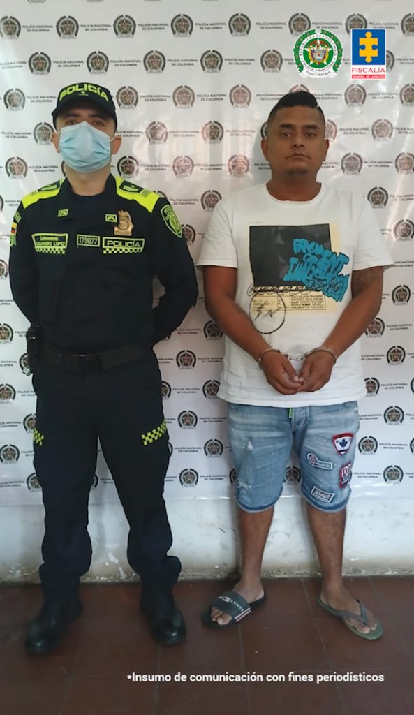 En la fotografía se aprecia de pie y con los brazos adelante a Diego Alexander Cortés Mera. Viste camiseta blanca de manga corta con estampado de colores negro, azul y amarillo en la parte frontal; bermuda de jean y pantuflas. Al lado derecho de él hay un uniformado de la Policía Nacional. Detrás de ellos hay un pendón institucional de la Policía.
