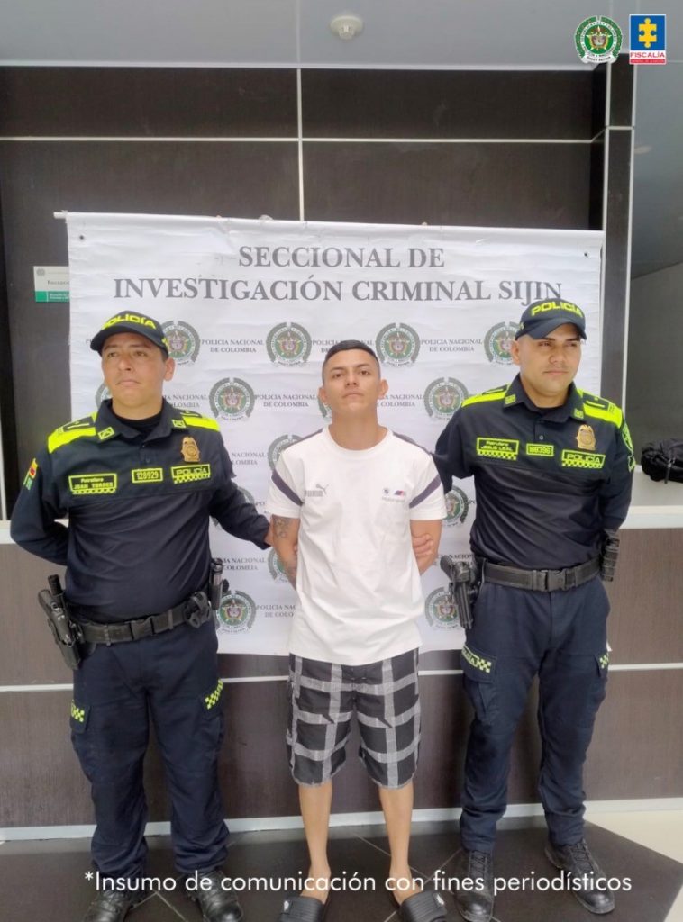 Se visualiza al capturado junto a dos uniformados de la Policía Nacional. Detrás de ellos el banner que identifica a la Seccional de Investigación Criminal -SIJIN de la Policía Nacional.