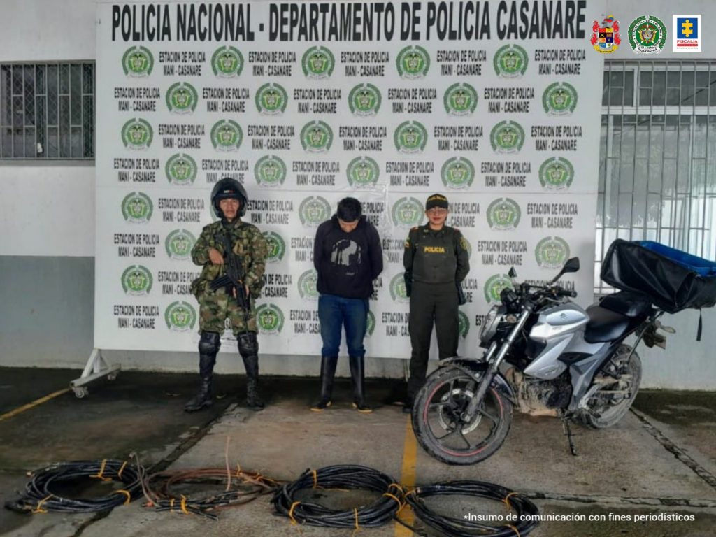 Está el capturado de frente esposado manos atrás, custodia por un militar y un policía. Delante de ellos la motocicleta y el cable de cobre incautado. Detrás banner de la Policía Nacional.