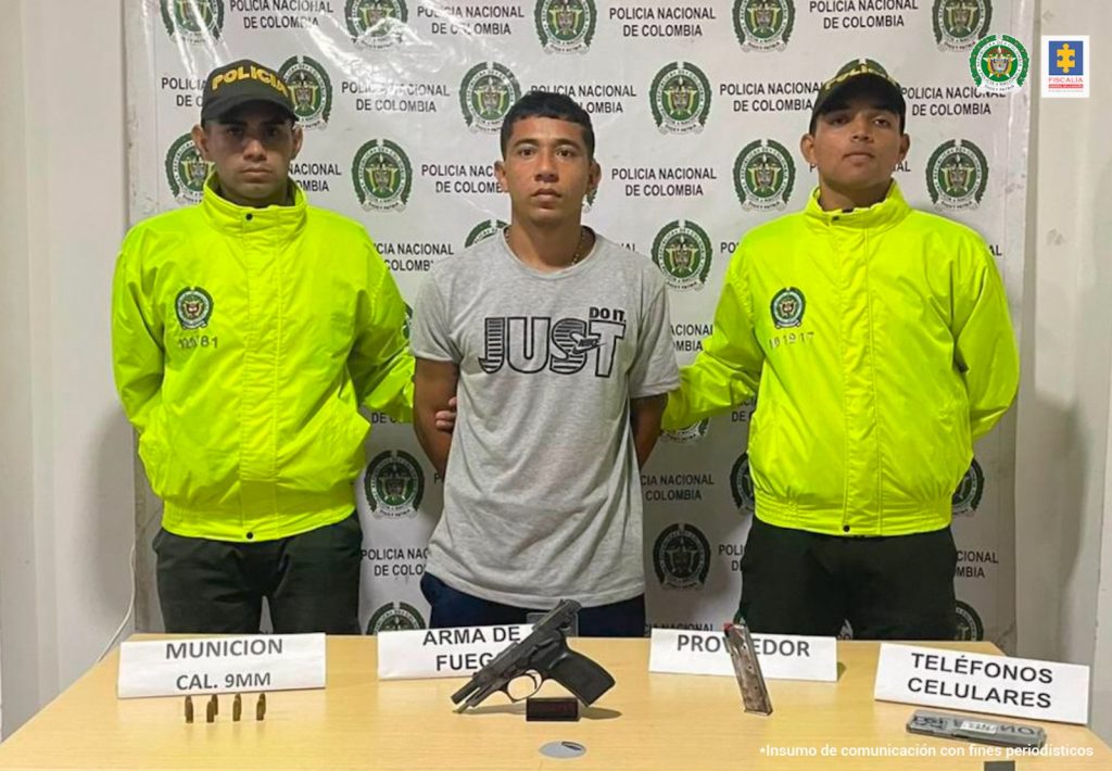 En la imagen se ve una persona detenida, entre dos Policías, y frente a una mesa que tiene un arma y munición.