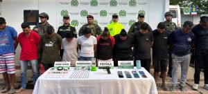 capturados de frente custodiados por uniformados de la Policía y del Ejército Nacional. Detrás banner de la Policía Nacional. Delante de los capturados mesa con material incautado.